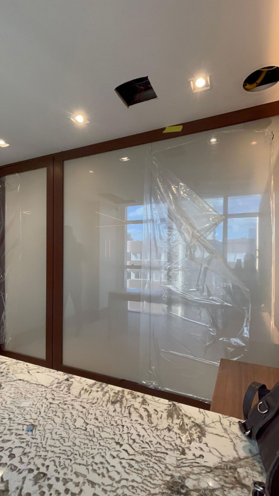 Divisória de vidro com película inteligente no modo privativo em ambiente corporativo em obra, vidro leitoso e opaco bloqueando totalmente a visão, plástico de proteção visível na parte superior, bancada de mármore em primeiro plano, fotografia realista