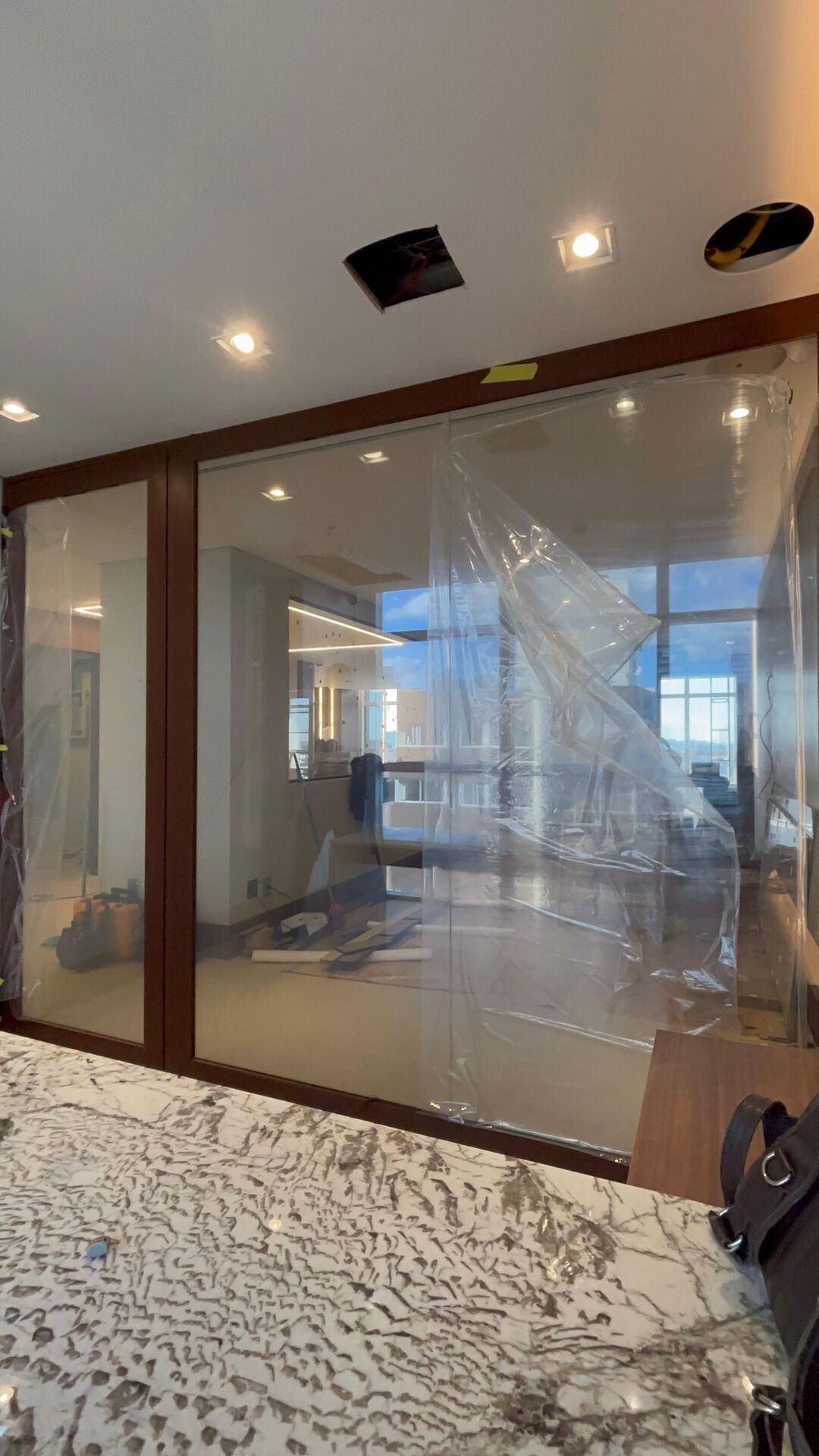 Divisória de vidro com película inteligente no modo transparente em ambiente corporativo em obra, vidro nítido revelando escritório ao fundo com bancada de mármore, plástico de proteção translúcido cobrindo parte da estrutura, tons neutros e madeira, fotografia realista